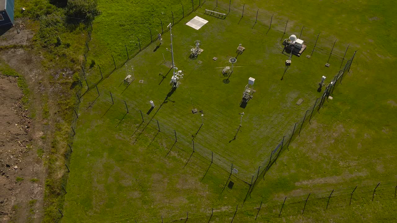 Aerial summer view of a fenced weather monitoring station with scientific instruments on a green field in Estonia captured on a sunny day
