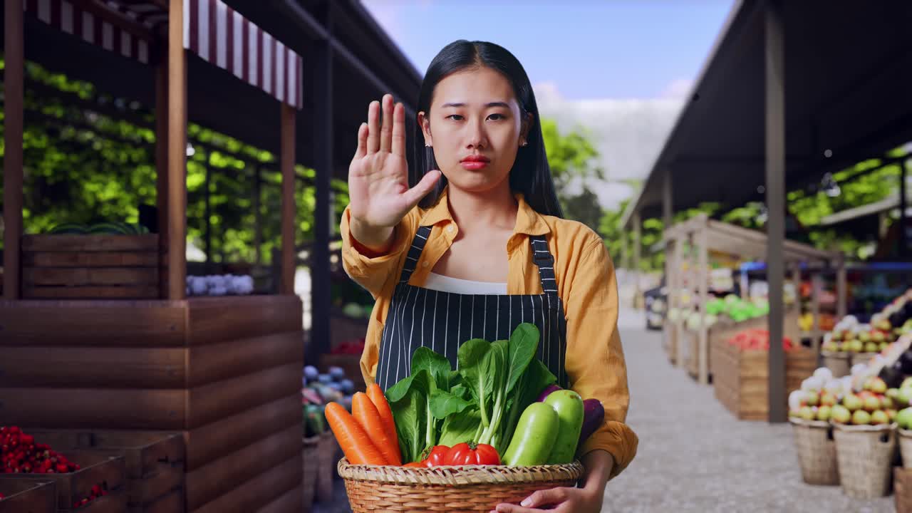 野菜かごを持ち、片手で制止のジェスチャーをしているアジア人女性農家、ファーマーズマーケットに立つ。