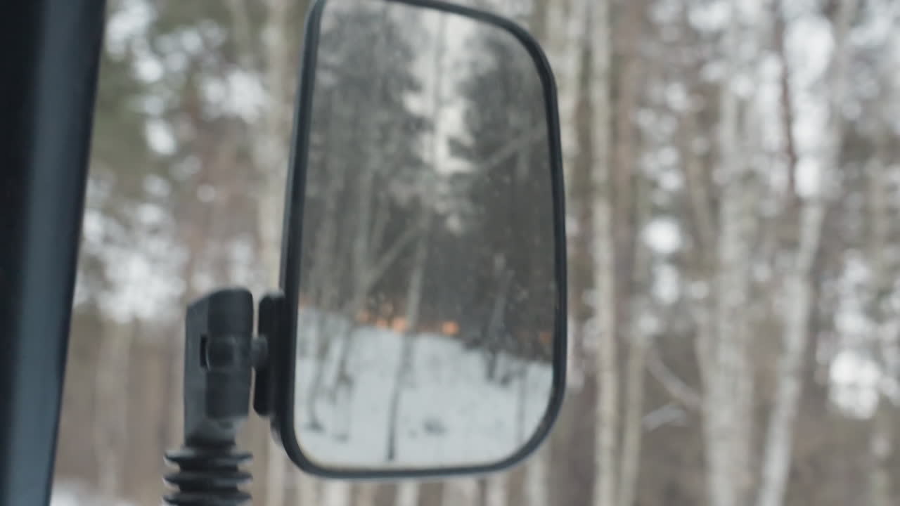 Side mirror reflecting snowy forest trail, frost and droplets on glass framing distant pines, mount and arm visible, rearview perspective captures quiet winter landscape and muted light while rider