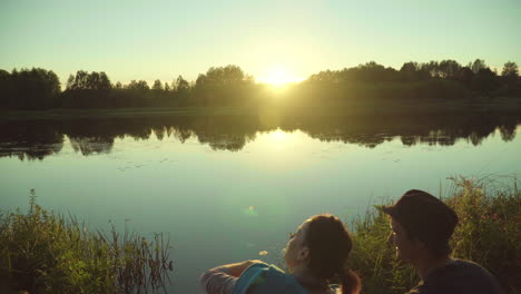 Los-Jóvenes-Disfrutan-De-Las-Vacaciones-Sentados-En-La-Orilla-Del-Río-Y-Contemplando-La-Puesta-De-Sol.-La-Gente-Se-Relaja