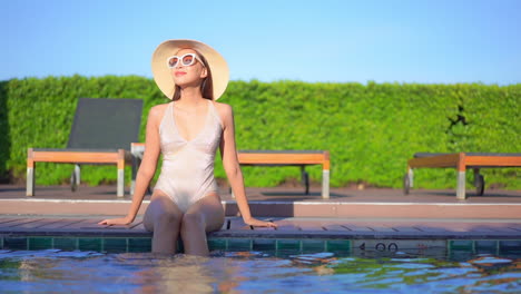 Mujer-Delgada-En-Traje-De-Baño-Sentada-En-El-Borde-De-La-Piscina-Disfrutando-Del-Sol-Vespertino-En-Un-Destino-Tropical