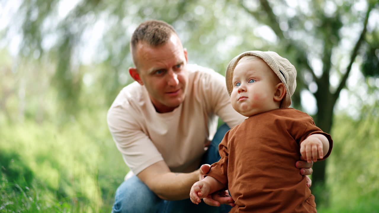 Cute chubby baby boy in a cap stands outdoors supported by dad. Daddy lets his son stand by himself and the kid sways a little.