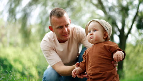 cute chubby baby boy in a cap stands outdoors supported by dad. daddy lets his son stand by himself and the kid sways a little.