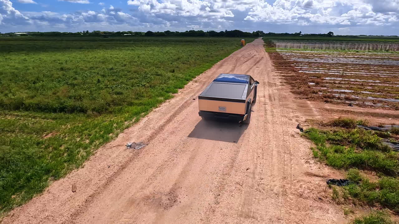 Cyberpunk-style electric car driving by the farm field. Tesla Cybertruck footage from FPV drone on sunny day.
