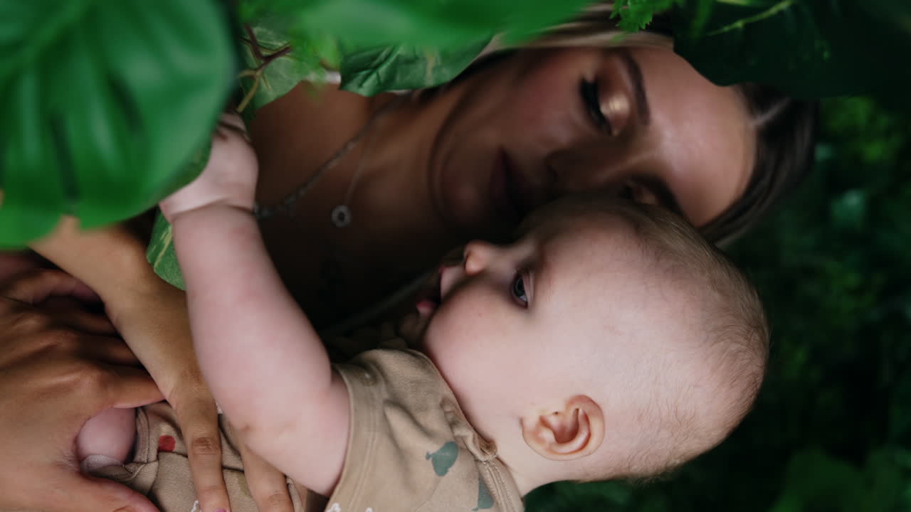 Woman with blonde long hair holds her baby in hands. Little infant is focused on the green leaves. Close up. Vertical screen.