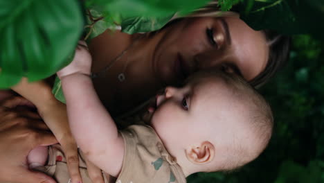 woman with blonde long hair holds her baby in hands. little infant is focused on the green leaves. close up. vertical screen.
