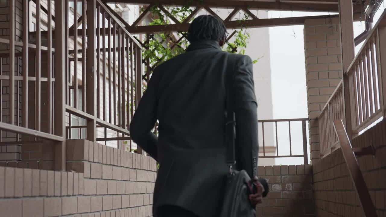 Back view of young adult in black suit rushes down hallway toward door, quick return to pick forgotten item inside house, decisive stride, indoor corridor lines, urgency and modern interior backdrop