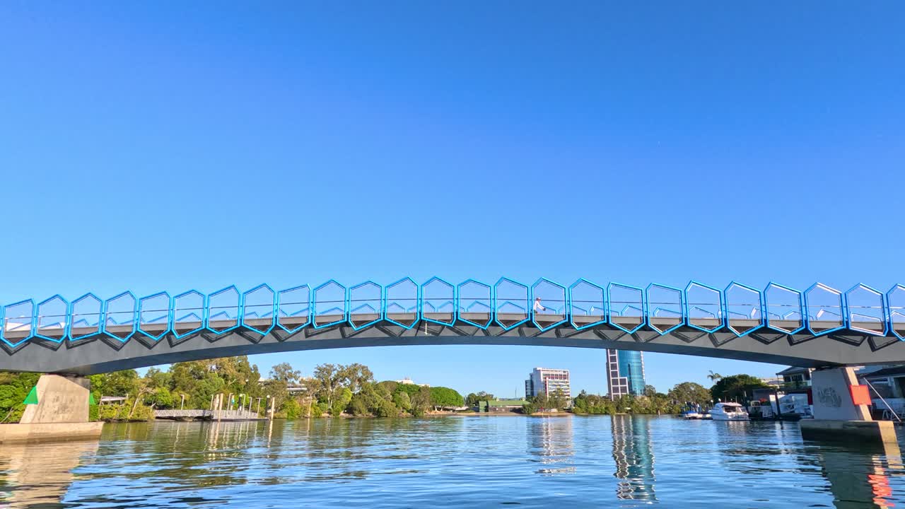 Camera glides smoothly beneath a contemporary pedestrian and bike bridge over the Nerang River, Gold Coast, in bright daylight with clear blue skies