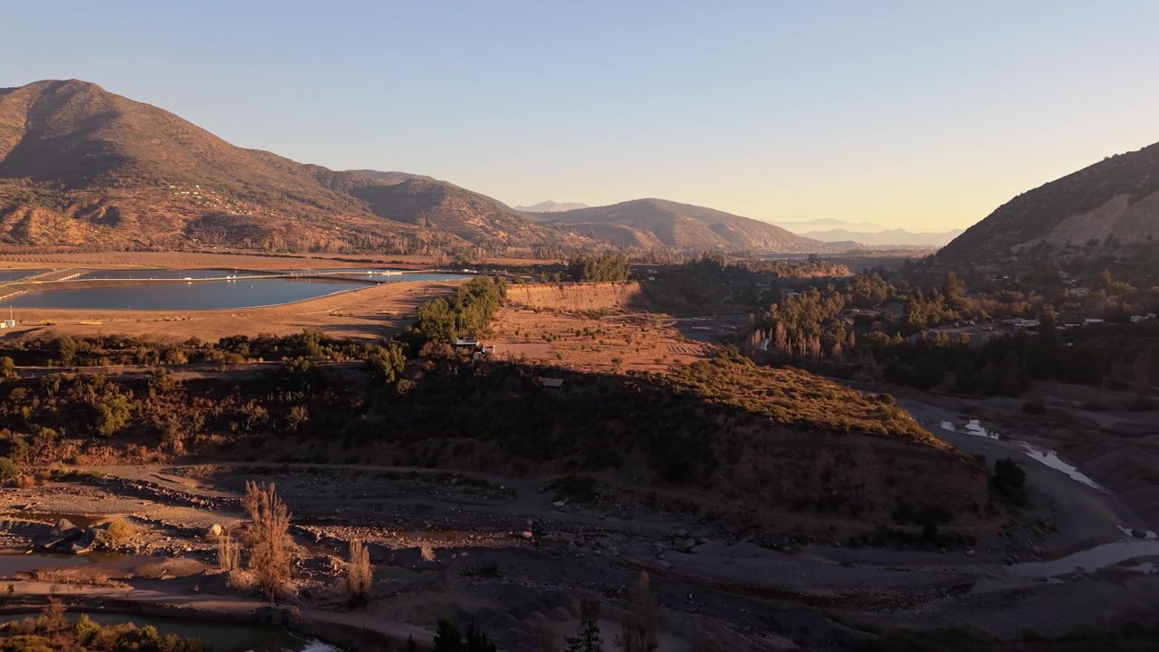 Volo all'indietro sopra un ampio letto di fiume roccioso e asciutto che rivela stagni geometrici di trattamento delle acque in lontananza, annidati tra le dolci colline pedemontane al tramonto.