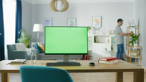 modern personal computer with mock-up green screen display standing on the desk of the cozy home office. in the background smart man takes book of the shelf of the bright living room.