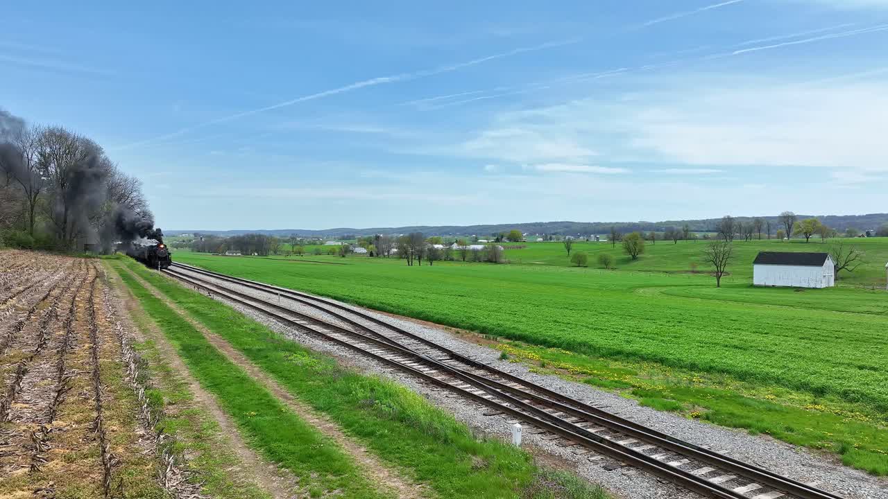 A vintage steam train moves along the tracks, surrounded by vibrant green fields and a white barn. The clear blue sky adds a serene atmosphere to the picturesque rural setting