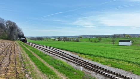 a vintage steam train moves along the tracks, surrounded by vibrant green fields and a white barn. the clear blue sky adds a serene atmosphere to the picturesque rural setting
