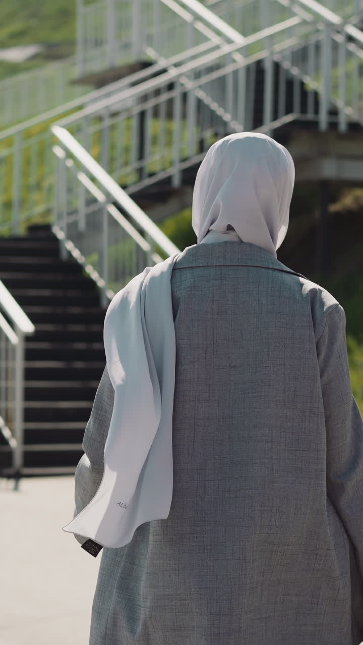 Arabian woman in grey coat adjusts hijab walking to stairs on hill slope backside view. Slim woman with Muslim headscarf against green hilly bank slow motion