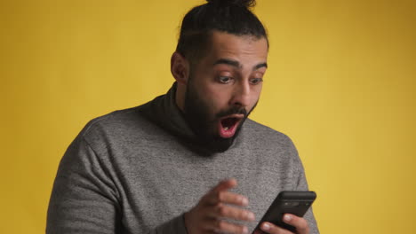 Foto-De-Estudio-De-Un-Hombre-Emocionado-Con-Un-Teléfono-Móvil-Celebrando-Ganar-Dinero-Contra-Un-Fondo-Amarillo