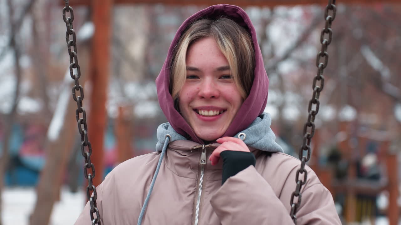 mujer joven sonriente con capucha sostiene collar de capucha mientras está sentada en el columpio en el parque de invierno, vestida con una chaqueta cálida, disfruta del clima frío con un fondo borroso de árboles y estructuras de patio de recreo