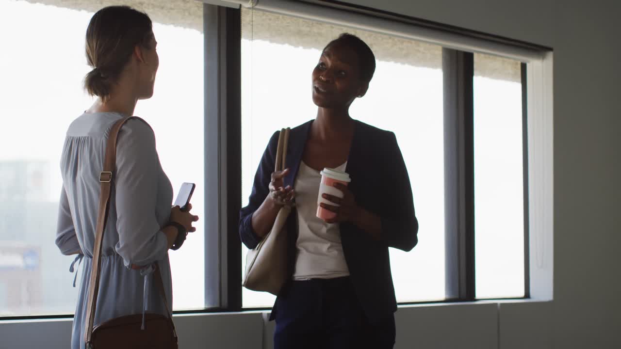 Two diverse female colleagues standing at window, talking and smiling