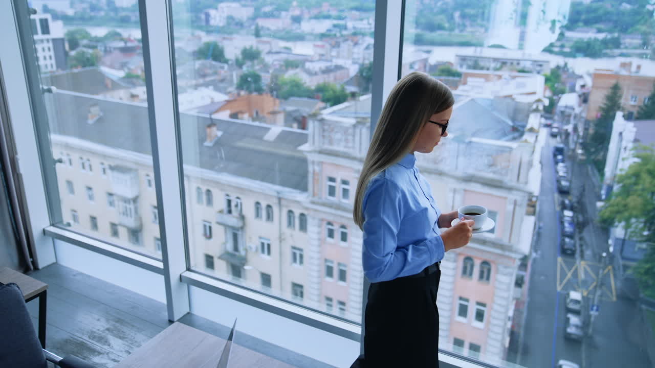 Lunch break in the modern office with beautiful view from window. Blonde female employee walking up to window with a cup of coffee. High angle view.