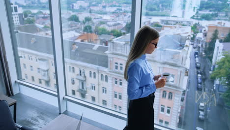 lunch break in the modern office with beautiful view from window. blonde female employee walking up to window with a cup of coffee. high angle view.