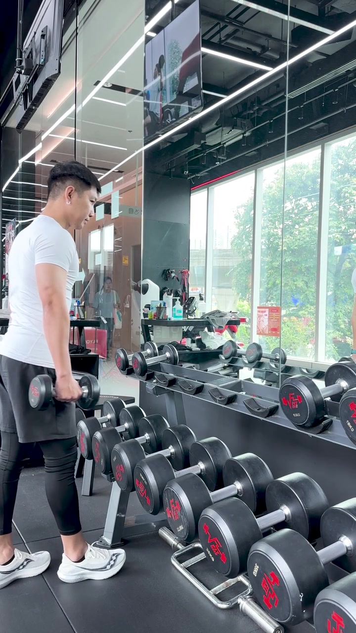 Dua pria memilih dumbel di gym yang terang, bersiap untuk latihan kekuatan dengan cahaya alami.