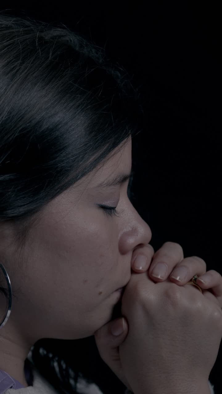 Thoughtful close-up shot captures a Hispanic woman praying with closed eyes in a vertical video format, perfect for mobile and social media. Intimate, serene atmosphere with dark background