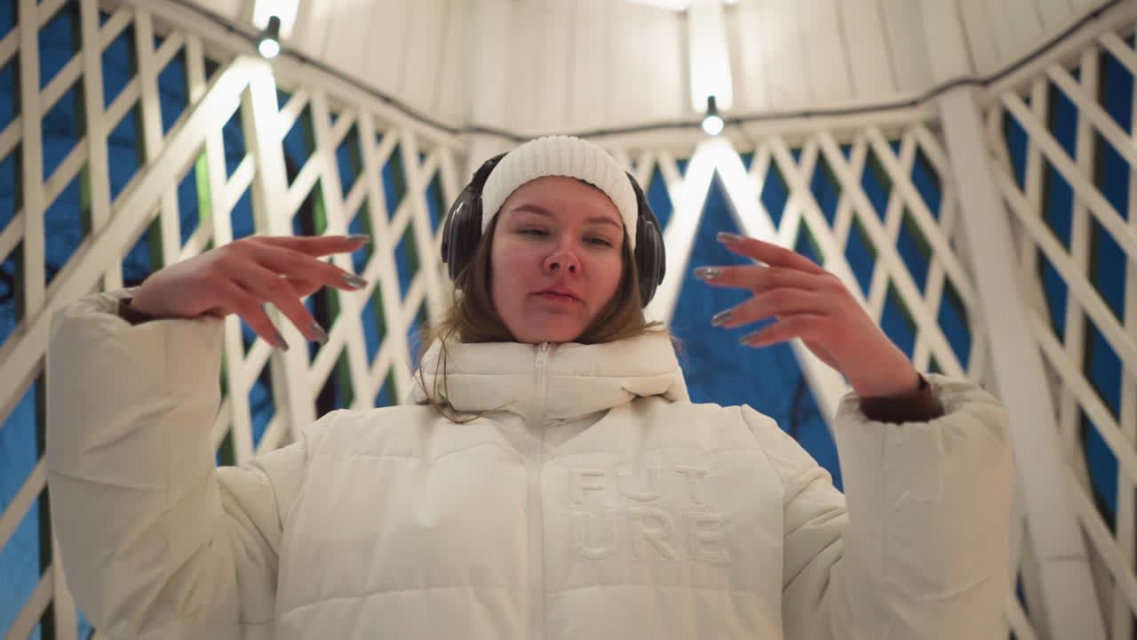 White lady wearing headphones closes eyes and moves hands passionately in rhythm of music inside illuminated latticed pavilion at dusk with snow softly blanketing floor and warm overhead lights