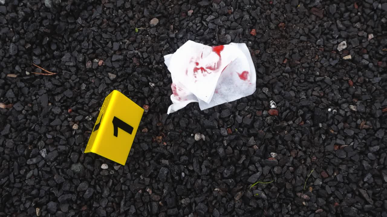 Vista dall'alto di un fazzoletto bianco sporco di sangue accanto a un segnale giallo della scientifica sulla scena del crimine, su un terreno roccioso scuro all'aperto, di giorno