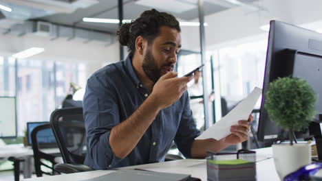 Mixed-race-businessman-sitting-at-desk-holding-document,-using-smart-phone-and-smiling