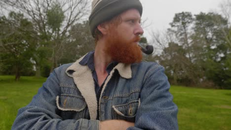 a shot of a bearded ginger mans face as he smokes from a tobacco pipe in the woods