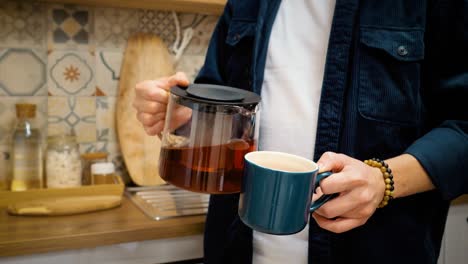 a man's hand pours tea into a mug. the man takes a sip and smiles at the camera. kitchen on the background