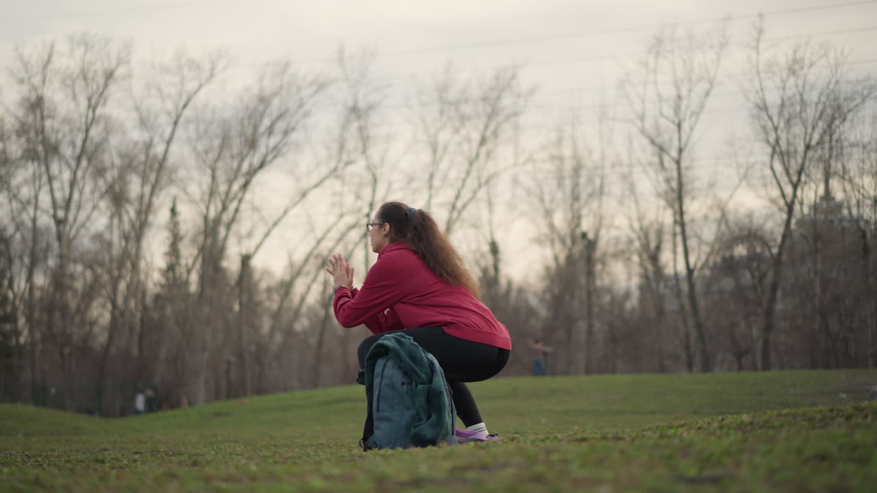 Young Female Lifting In Grassy Urban Setting, Lady Performing Lower Body Exercises Amidst City Park Scenery, Young Woman Engages In Squat Workout Under Cloudy Sky Near City Trees And Backpack