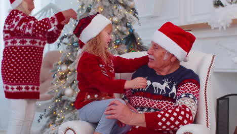 Granddaughter-child-fixes-Christmas-Santa-Claus-hat-on-happy-senior-old-smiling-grandfather-at-home