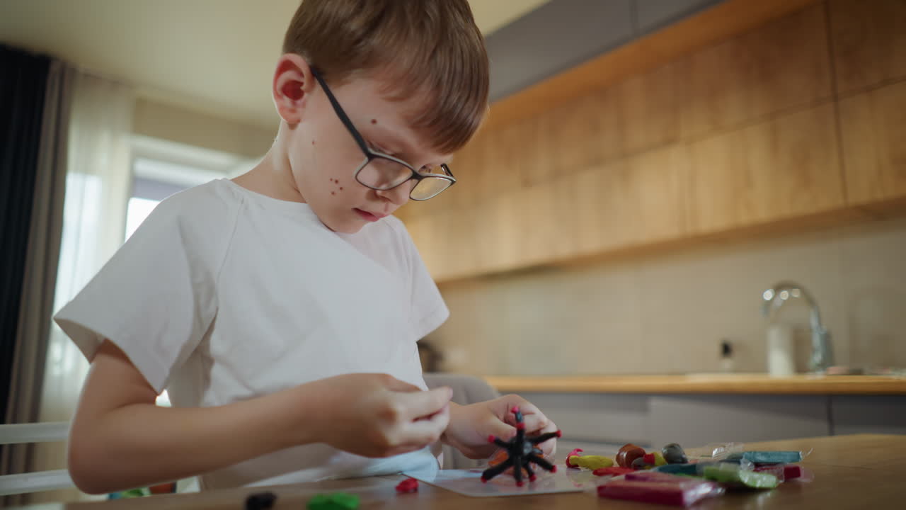 Ragazzo con gli occhiali concentrato a scolpire una figura di argilla a un tavolo di legno in cucina, circondato da argilla colorata per modellare, che esprime creatività, attenzione e apprendimento al chiuso