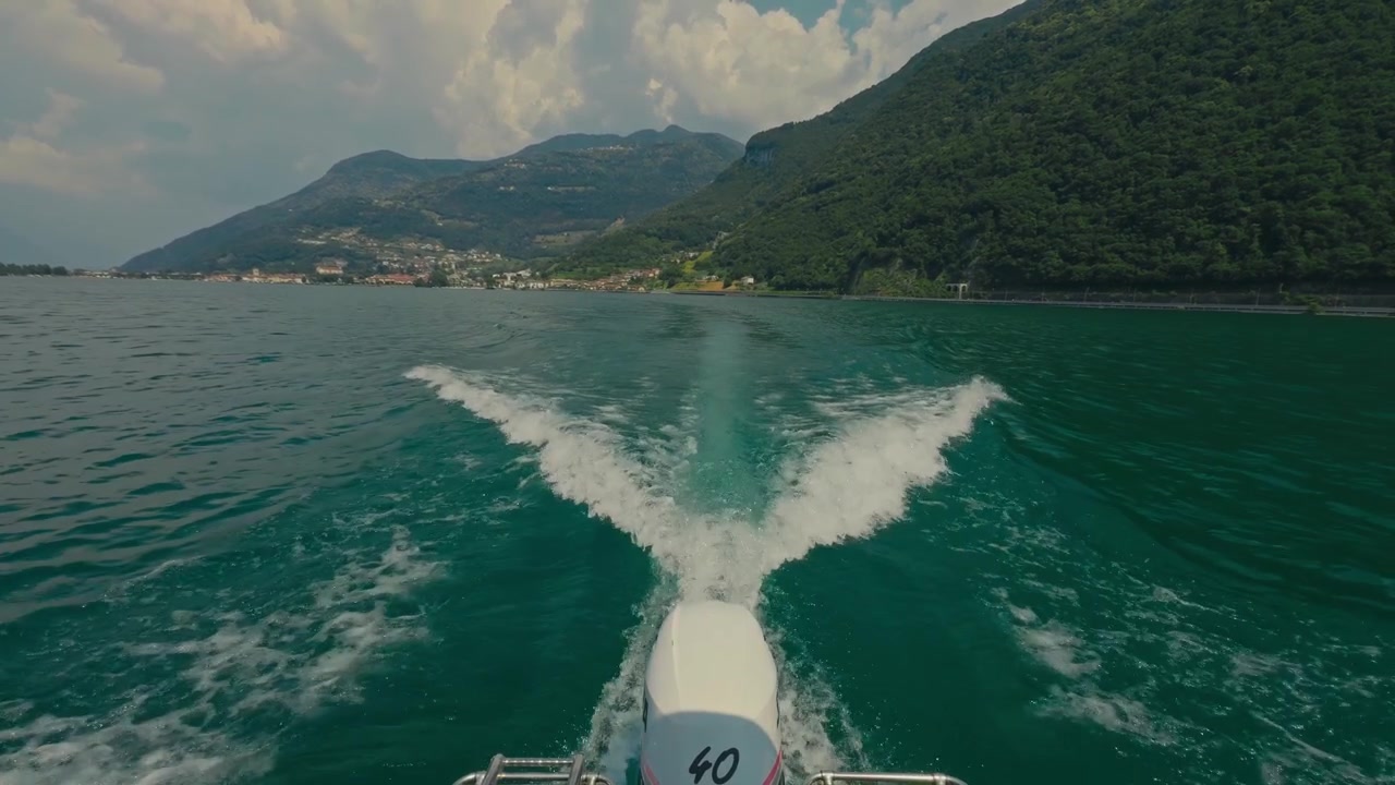 POV (punto di vista) dal ponte di una barca che guarda la scia spumeggiante attraverso il cristallino Lago d'Iseo, circondato da lussureggianti montagne verdi sotto un cielo estivo azzurro brillante nel nord Italia