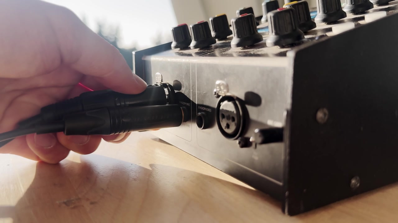 Nærbillede af en hånd, der trækker et kabel ud fra en broadcast intercom-lydenhed, med kontrolknapper, headset-stik, sollys og detaljer fra live produktionsudstyr.