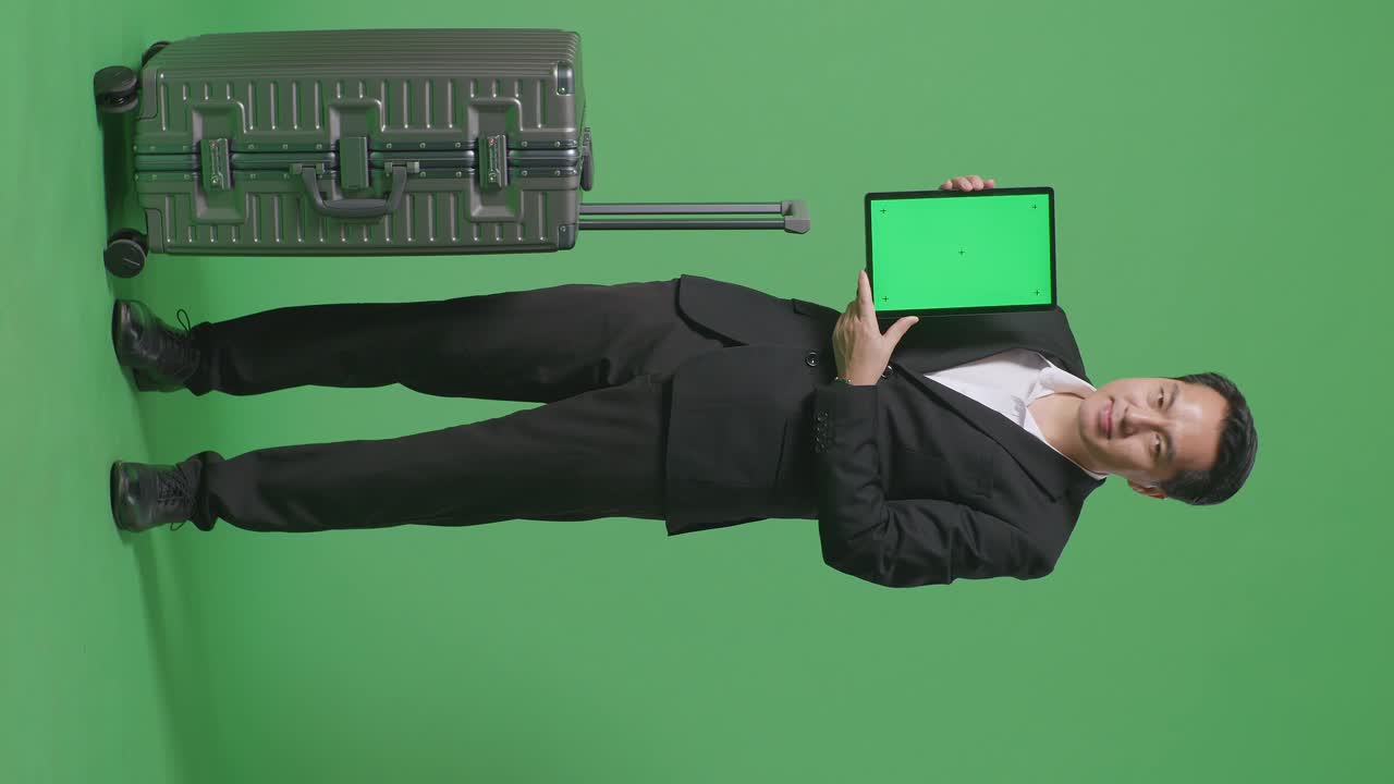 cuerpo lleno de viajero hombre de negocios asiático con equipaje sonriendo y mostrando la tableta de pantalla verde a la cámara mientras estaba de pie en el estudio de fondo de pantalla verde