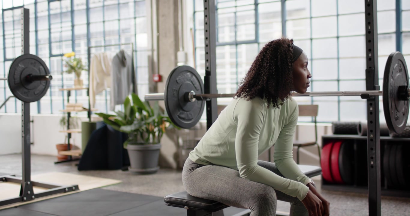 Donna afroamericana seduta su una panca in palestra con leggings verdi che si prepara per una serie con il bilanciere.