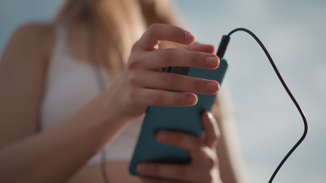 Primer plano de un teléfono en manos de una mujer caucásica en el exterior ajustándose unos auriculares con cable mientras consulta una lista de reproducción. Momento de calma y preparación antes del ensayo en la calle, con luz solar suave y poca profundidad de campo, con los dedos enfocados, en un ambiente moderno e informal.
