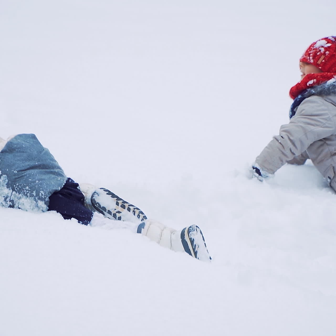 Back view of two kids crawling on snow on the white background in winter. Happy children are lying in soft snow outdoors. Close- up. Slow motion.