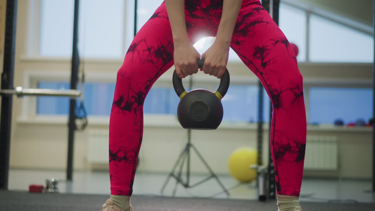 Atleta donna in palestra solleva un kettlebell, con postura sumo ampia, leggings luminosi, spinta mirata della parte inferiore del corpo, discesa controllata e potente hip thrust, atmosfera da dimostrazione di allenatore con attrezzatura da studio.