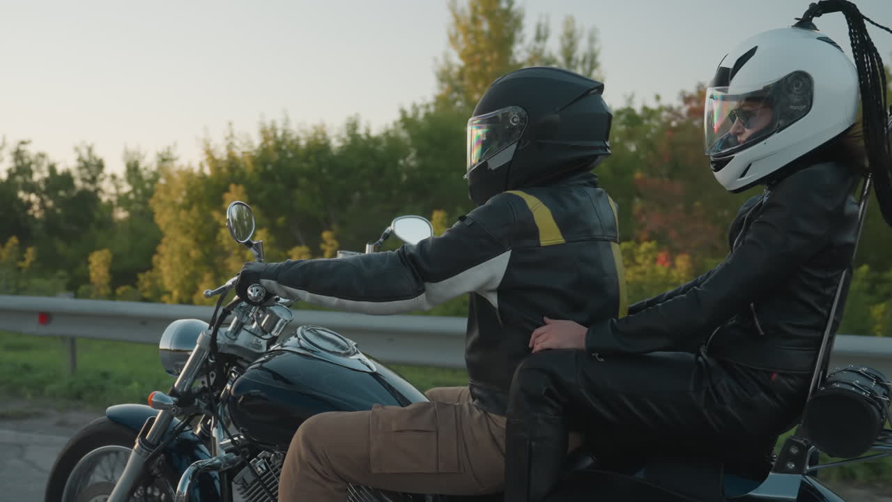 Couples traveling on power bike with chrome details and leather gear during sunset ride, front wheel and engine in focus, reflecting light while moving on asphalt road