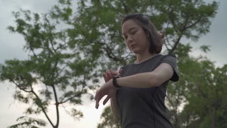 asian athlete woman using sport smart watch green nature park in evening sunset. female in sportswear touching screen application tracking activity and music. enjoy personal record data performance.