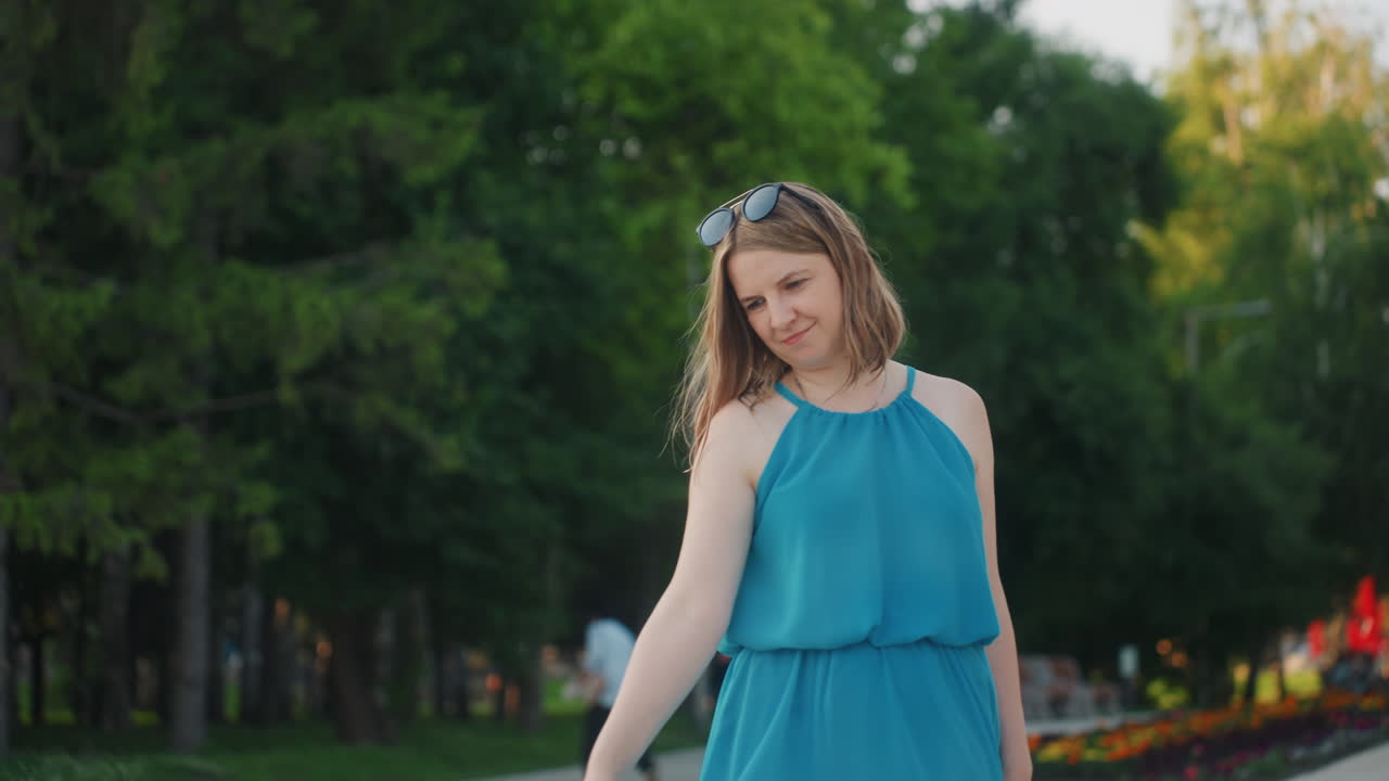 Fulllength caucasian woman in blue dress twirling on park pathway at golden hour, flowing skirt and sunglasses on head, playful pose as model during relaxed weekend outing, trees and flowerbeds