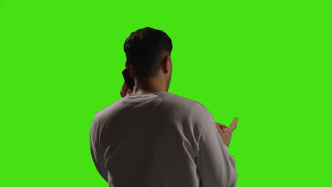 vista trasera de un joven hablando en el teléfono móvil de pie contra el fondo del estudio de pantalla verde con iluminación de baja clave