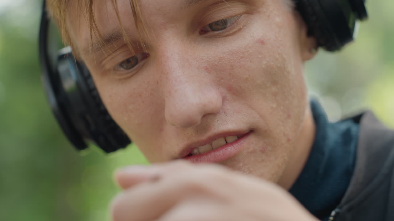 Un uomo bianco si concentra su una piccola formica con le cuffie, studio ravvicinato delle dita e dell'espressione. L'ascoltatore guarda intensamente la punta del dito, atmosfera contemplativa in un parco, accenni a un podcaster o a un hobby