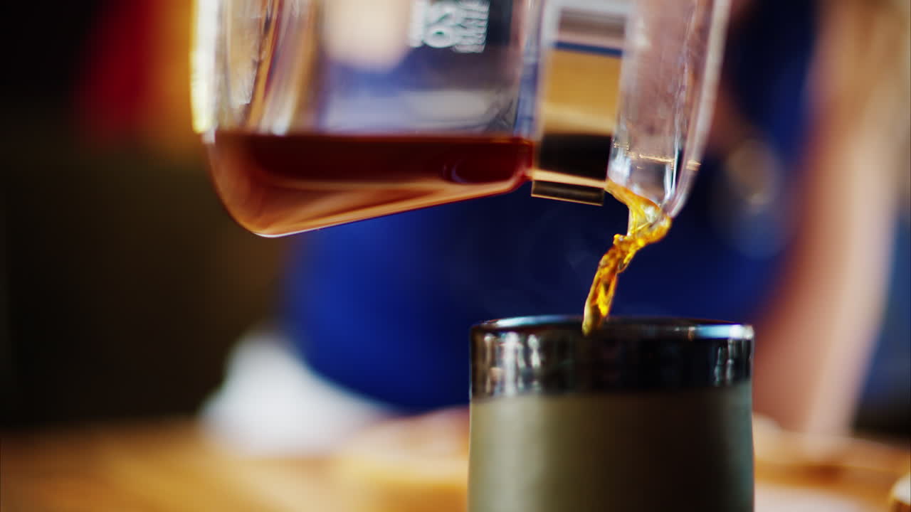 Close up of a woman pouring coffee in a cup from a glass pot standing on a wooden tray
