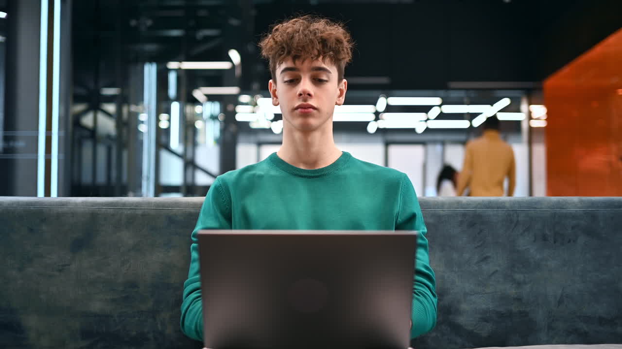 A young man working using a laptop while sitting on a sofa in an office