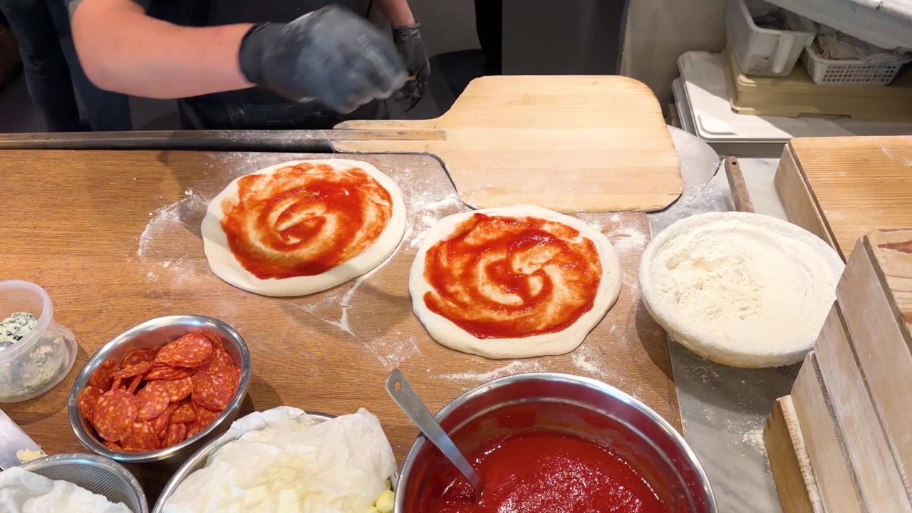Ripresa dall'alto di uno chef con guanti neri che condisce l'impasto della pizza con salsa di pomodoro e farina.