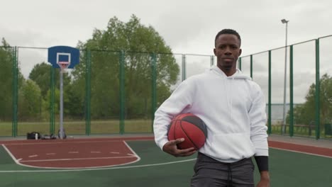 tracking portrait shot of young black man in hoodie standing on outdoor basketball court and holding ball