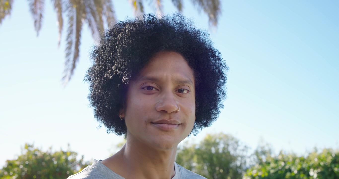 Afroameričan na dvoře u palmových listů, dívá se, jak se mění sluneční záře, sklání pohled, usmívá se.