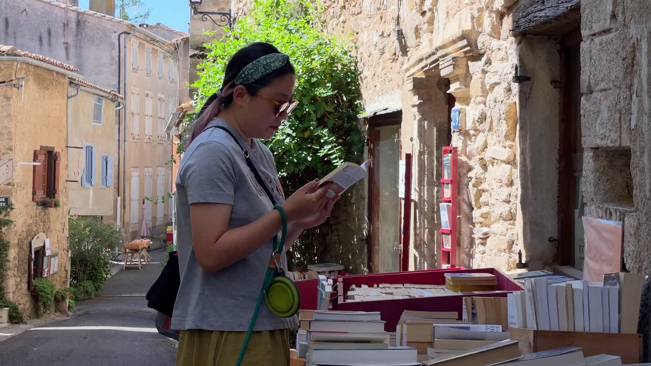 en la entrada de una librería de segunda mano con paredes moteadas en un pequeño pueblo de francia, una mujer asiática está hojeando libros